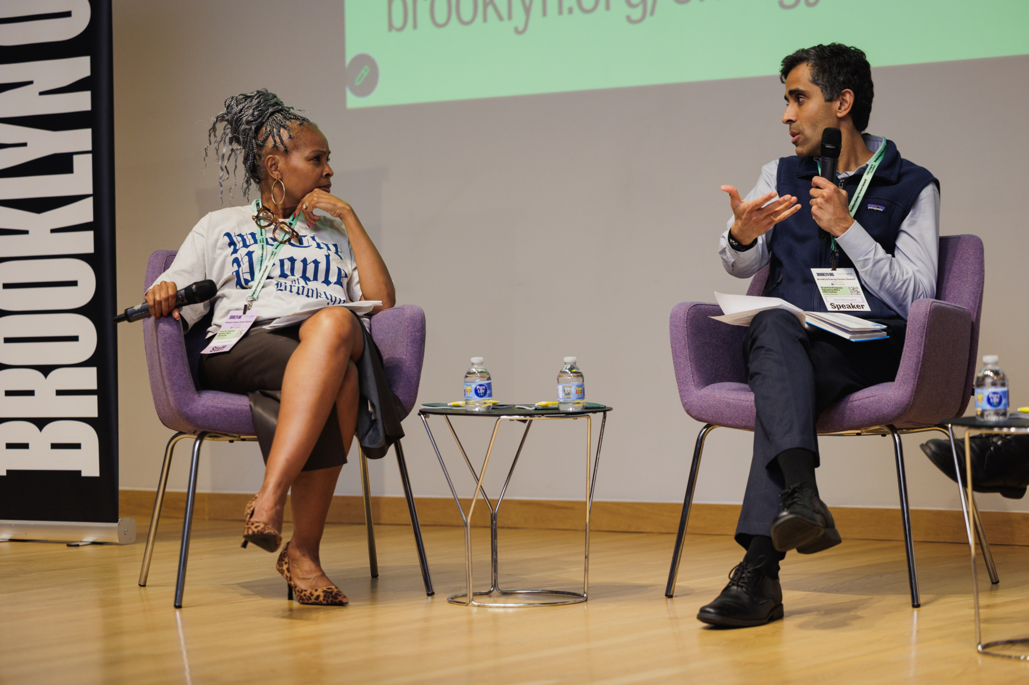 Two speakers sit in purple chairs on a stage, facing each other during a panel discussion.