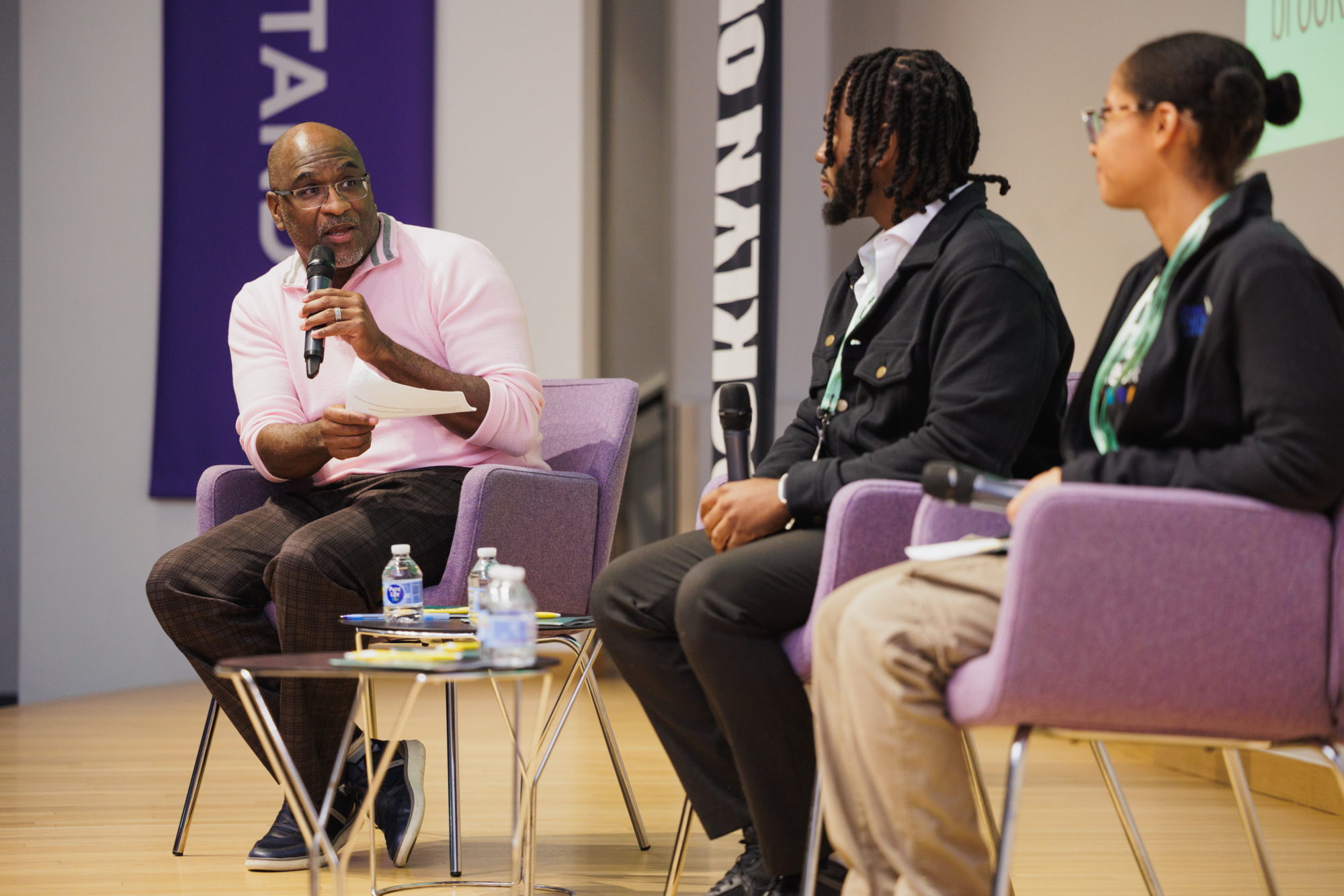 Three people sitting in armchairs on a stage, with the person on the left speaking into a microphone.