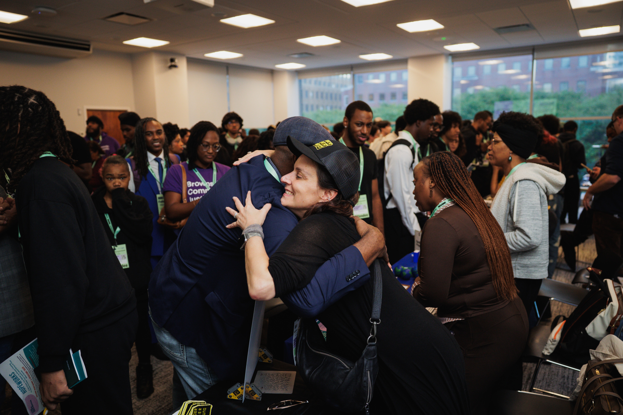 A woman in a black shirt and cap hugs a man in a blue blazer at a crowded indoor event.