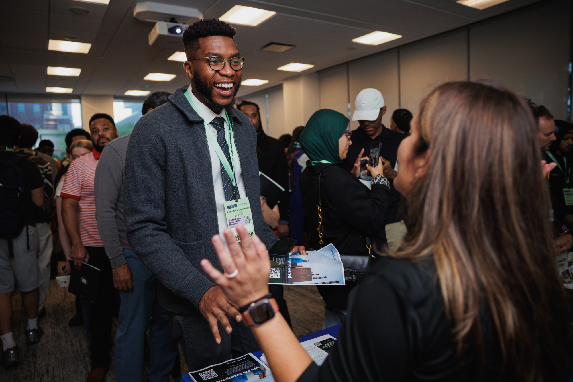 A man in a grey sweater and tie smiles while talking to a woman at a career fair, with other attendees in the background.