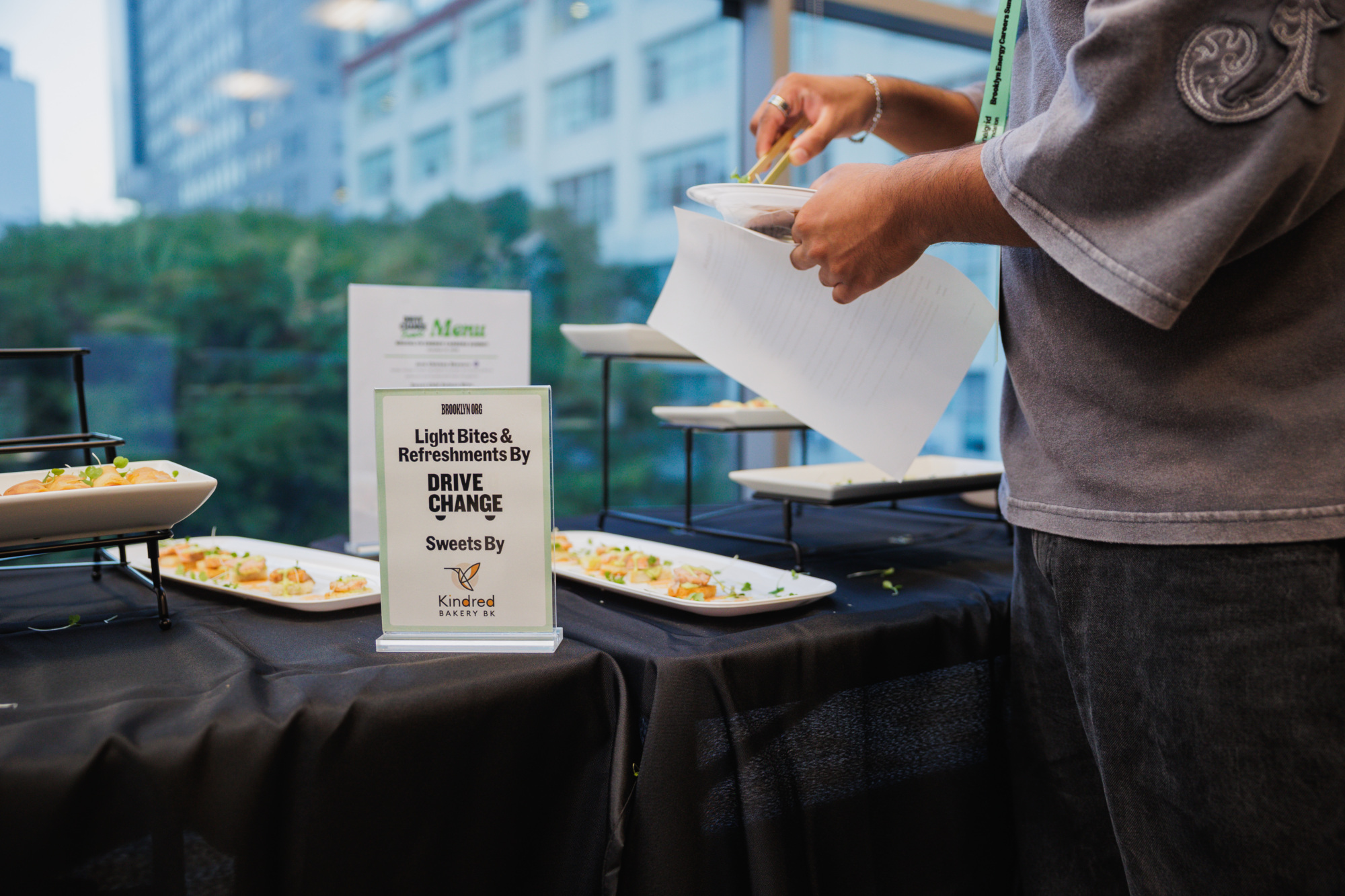 A person reaches for a small hors d’oeuvre from a serving tray near a sign that reads “Light Bites & Refreshments By DRIVE CHANGE.”