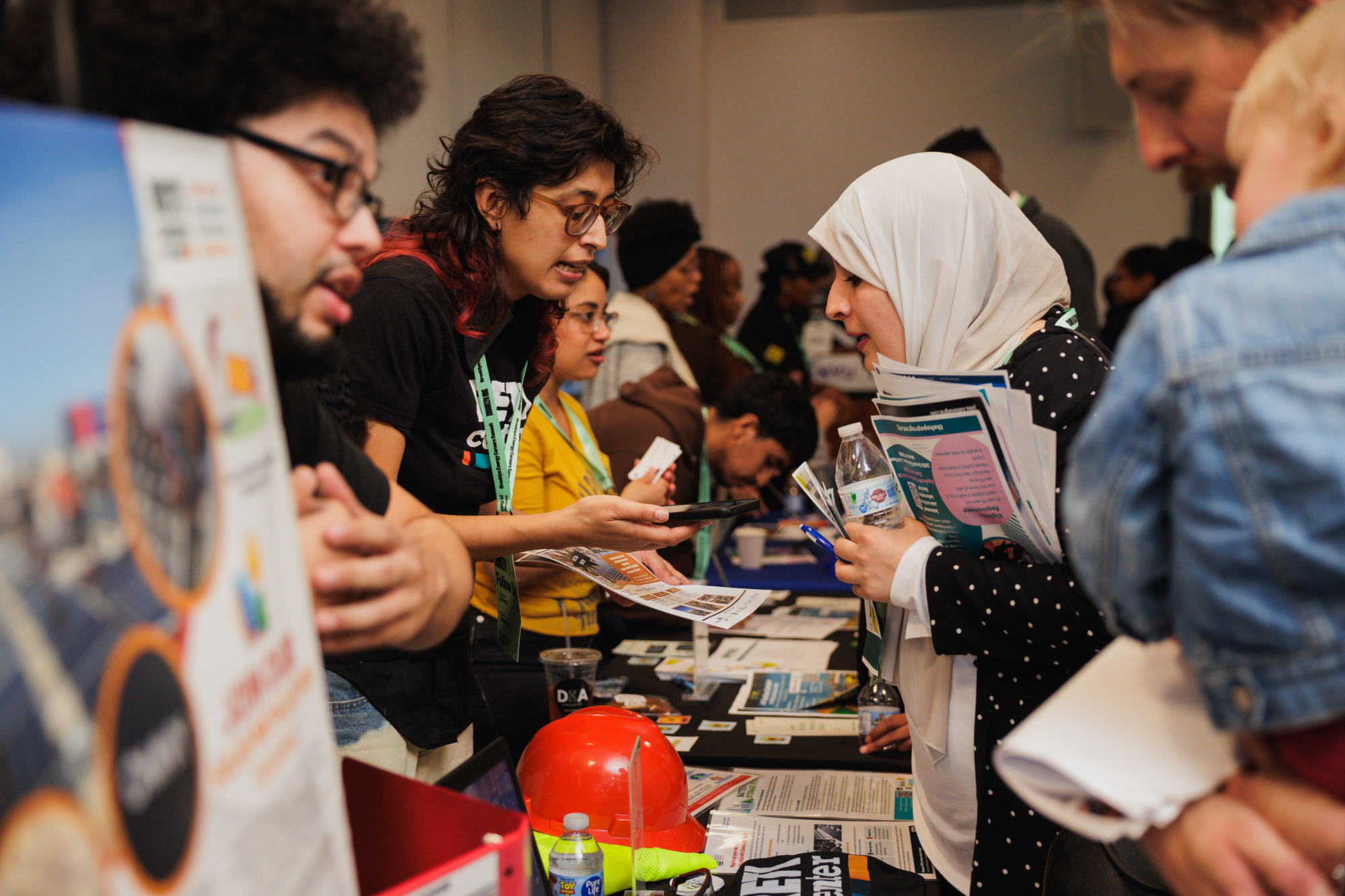A woman in a hijab speaks with a booth representative at an information fair while holding a stack of brochures.