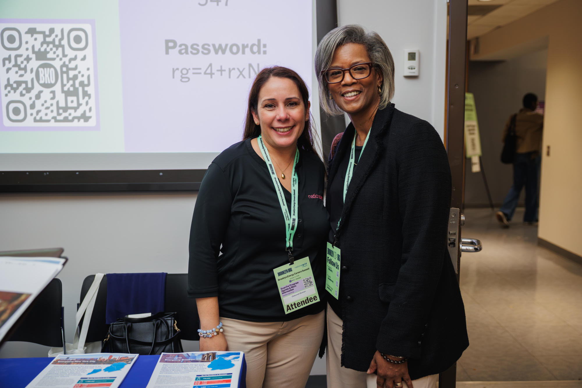 Two women wearing event lanyards stand in front of a projection screen displaying a QR code and password.