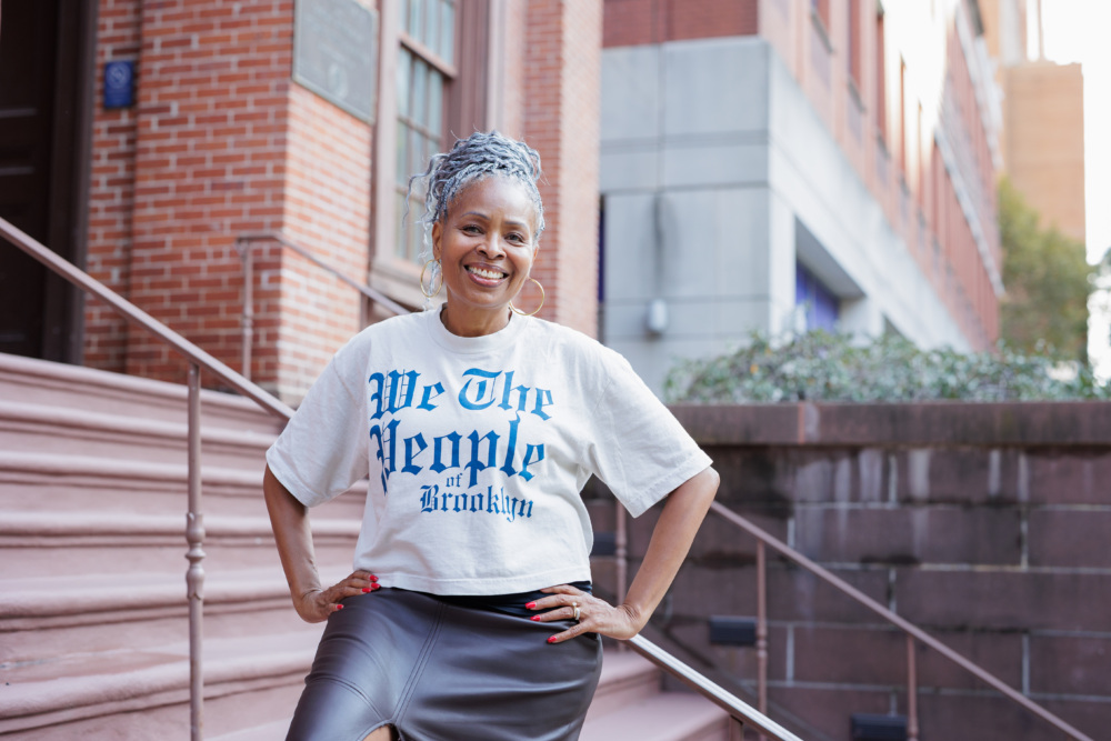 A smiling woman with grey hair wearing a white t-shirt that says “We The People of Brooklyn” stands on a brick building’s front steps with her hands on her hips.