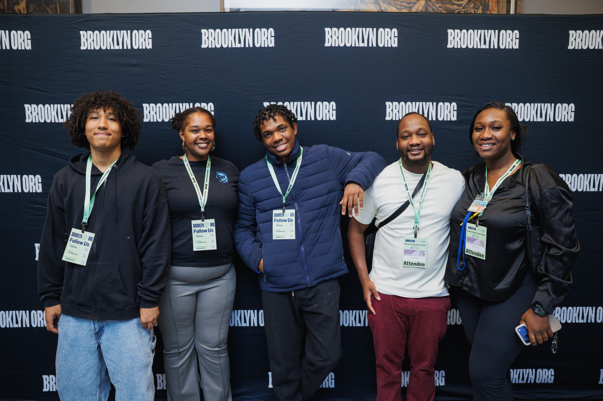 Five people stand in a row in front of a black step-and-repeat banner that reads “BROOKLYN ORG.” Each person is wearing a branded event lanyard and name badge.