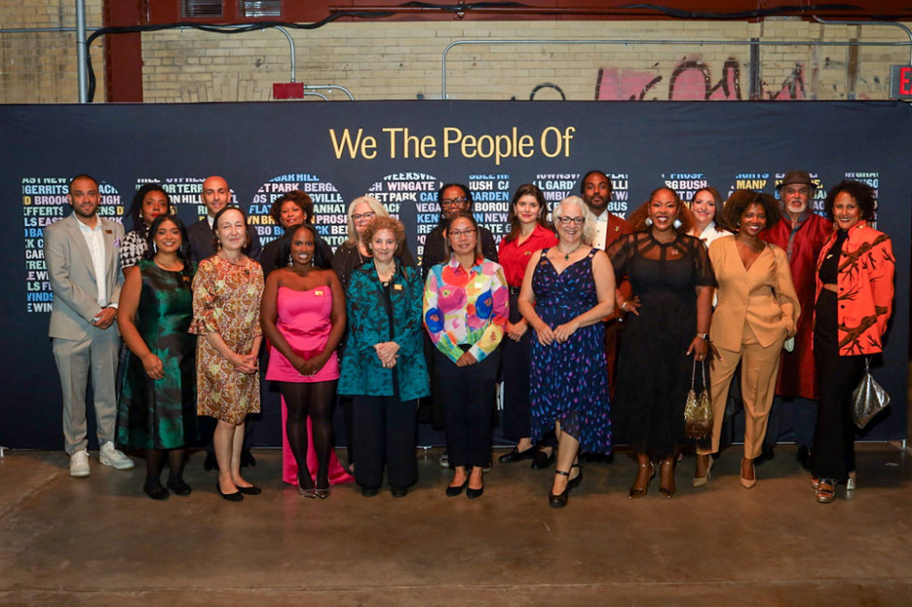 A group of nineteen people posing for a photo in front of a blue backdrop with the text “We The People Of” above a list of neighborhood names.