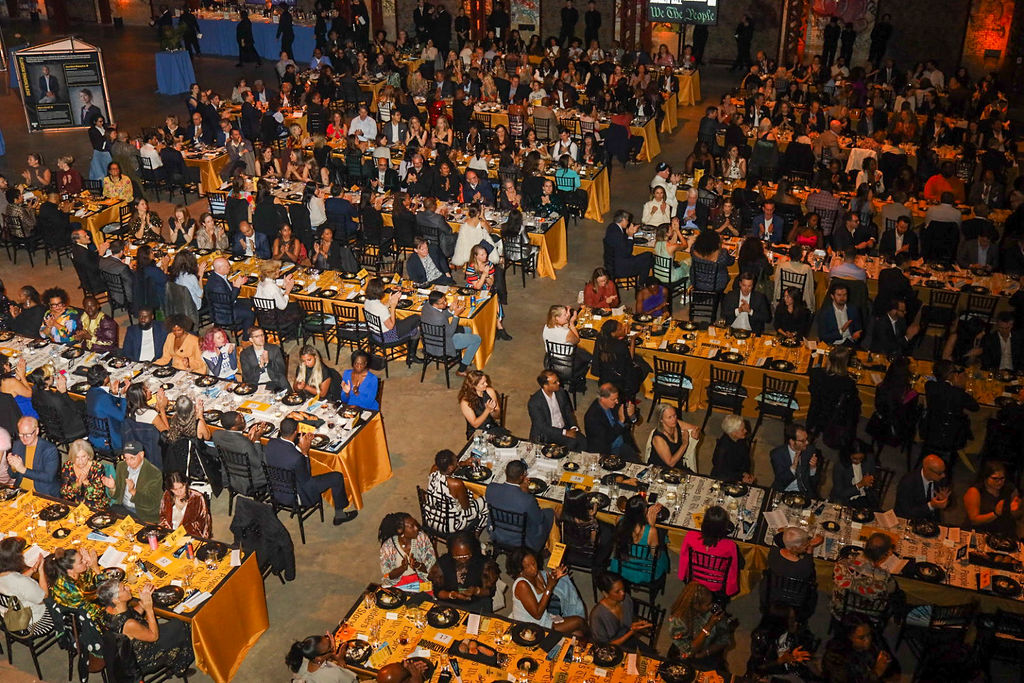 A high-angle view of a crowded gala or conference dinner with many people seated at round tables covered in gold tablecloths.