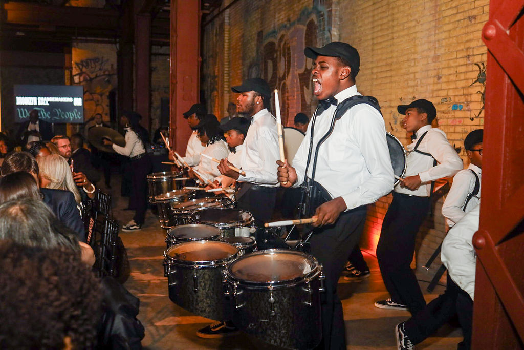 A line of young drummers in white shirts and black caps perform in a brick-walled room.