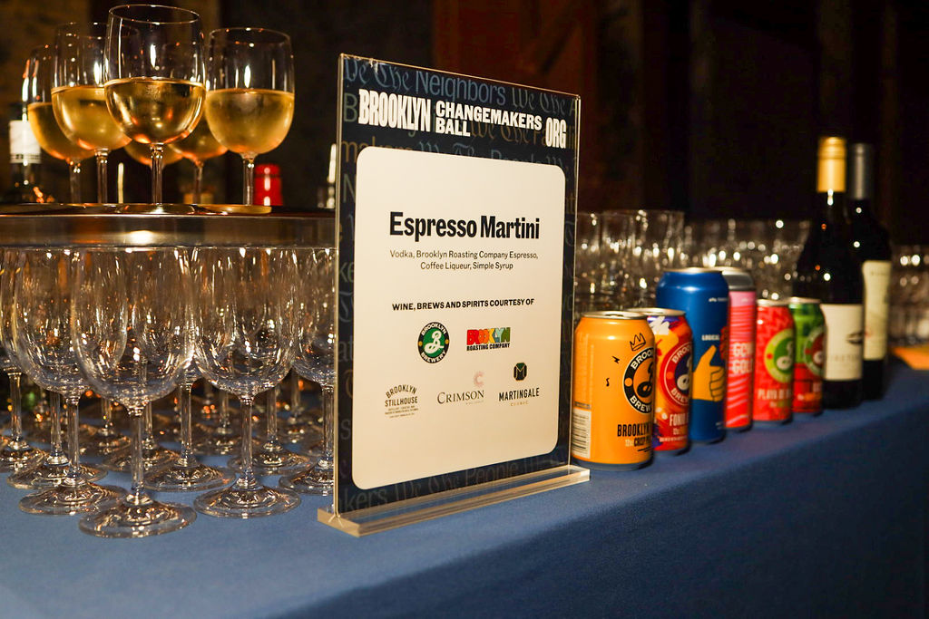Table sign for an Espresso Martini at the Brooklyn Changemakers Ball, surrounded by wine glasses and assorted cans of beer.