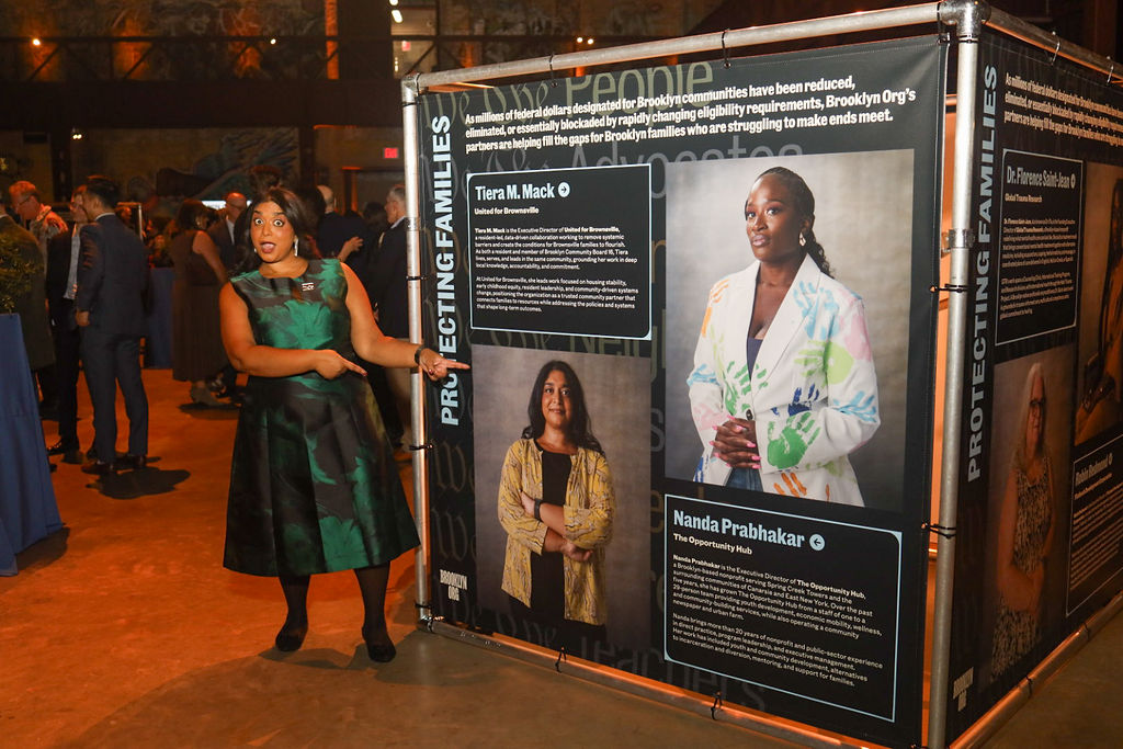 A woman in a green dress gestures toward a display banner featuring photos and biographies of community leaders.