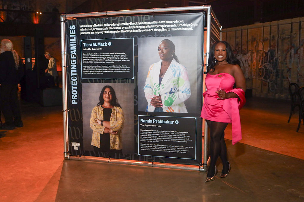 A woman in a pink dress stands next to a large display board featuring portraits and biographies of two community leaders.