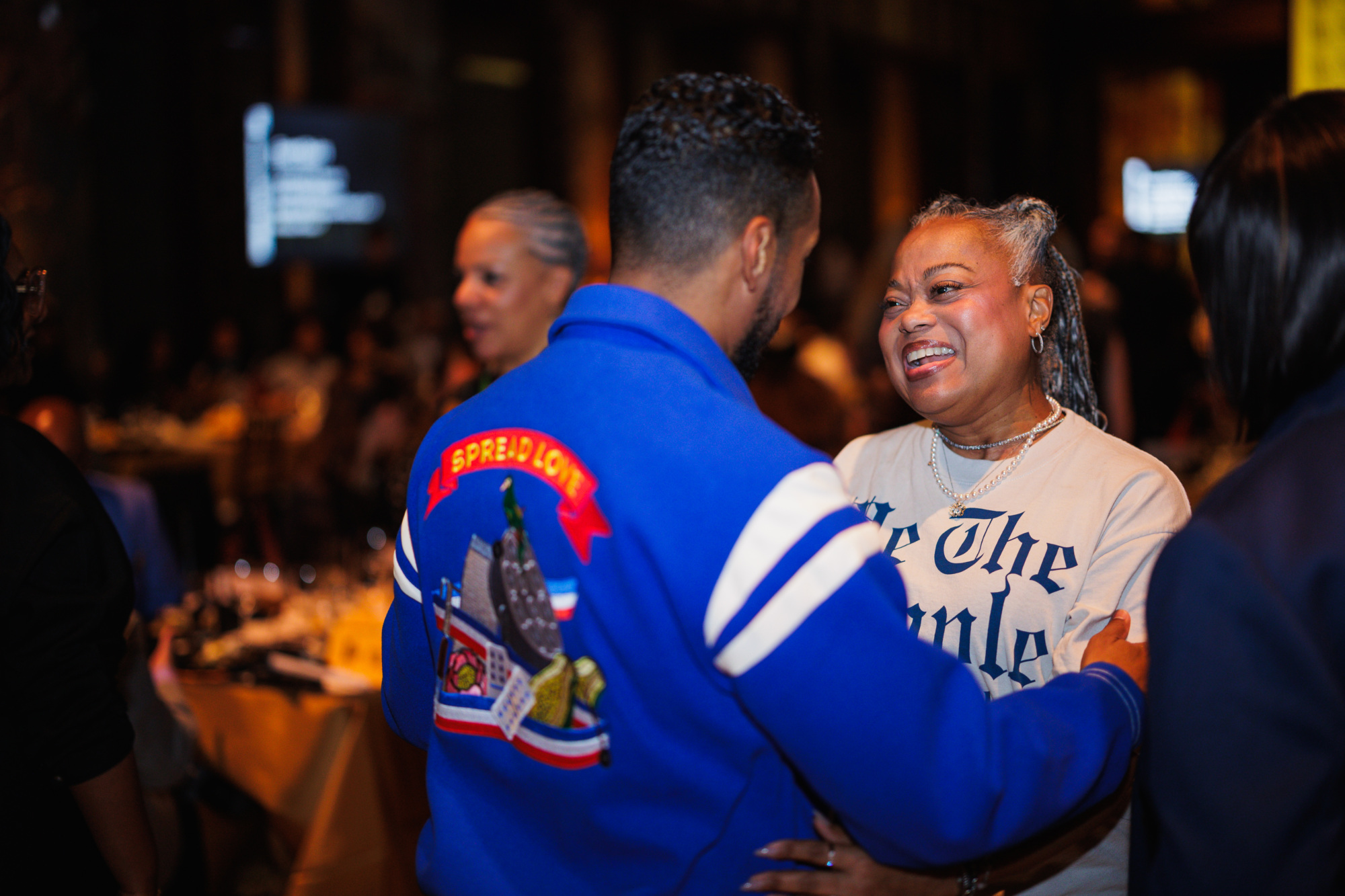 A man in a blue jacket with “SPREAD LOVE” on the back hugs a smiling woman wearing a “Be The People” shirt.