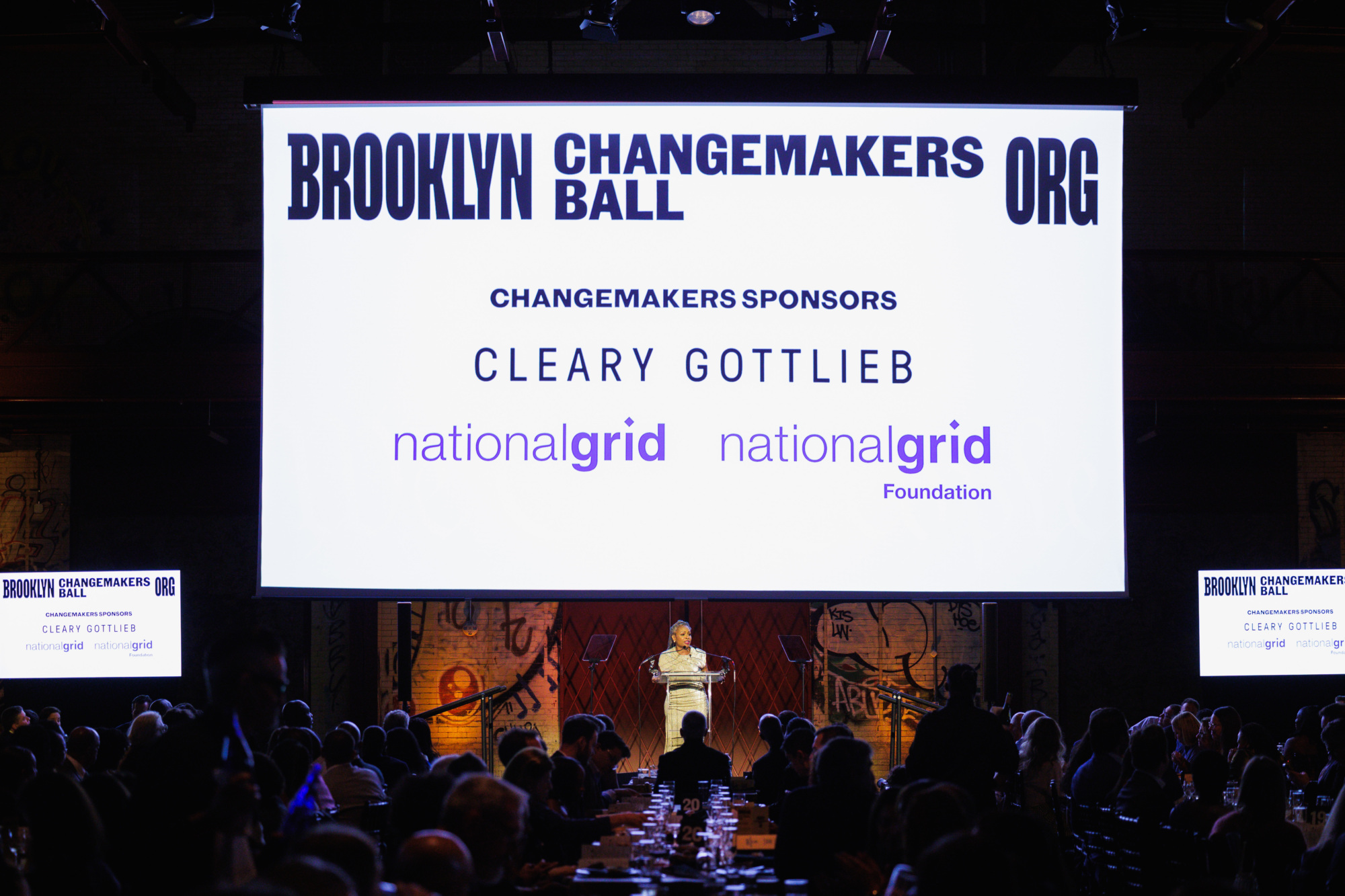 A speaker stands at a podium in front of a large screen displaying the “Brooklyn Changemakers Ball” logo and sponsor names: Cleary Gottlieb, National Grid, and National Grid Foundation.