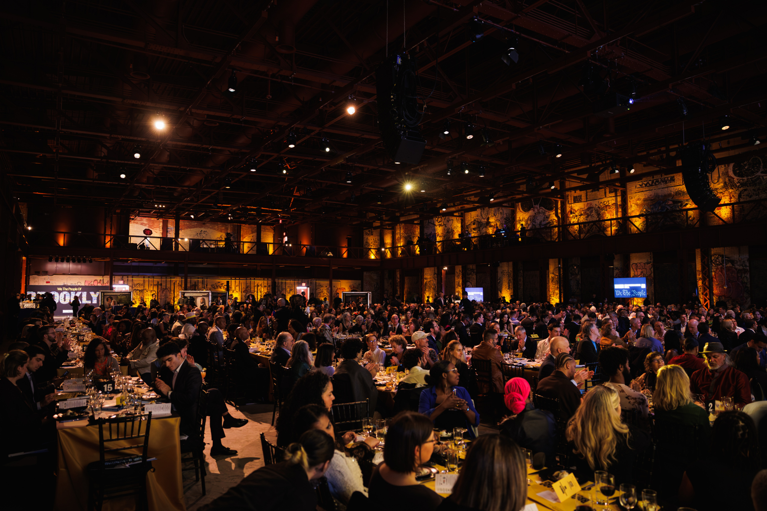 A large crowd of people seated at tables in a dimly lit, industrial event space.