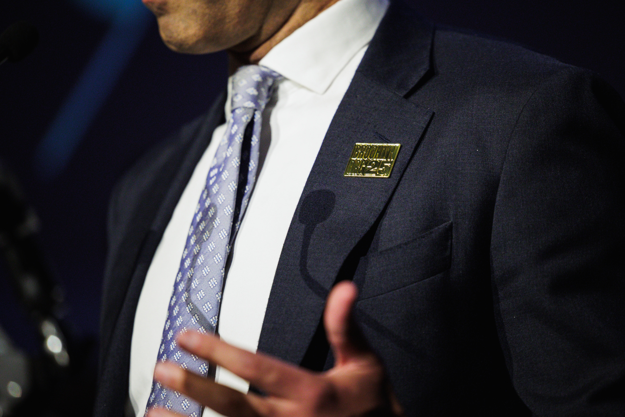 A person in a suit and tie with a gold “Brooklyn 175” lapel pin.