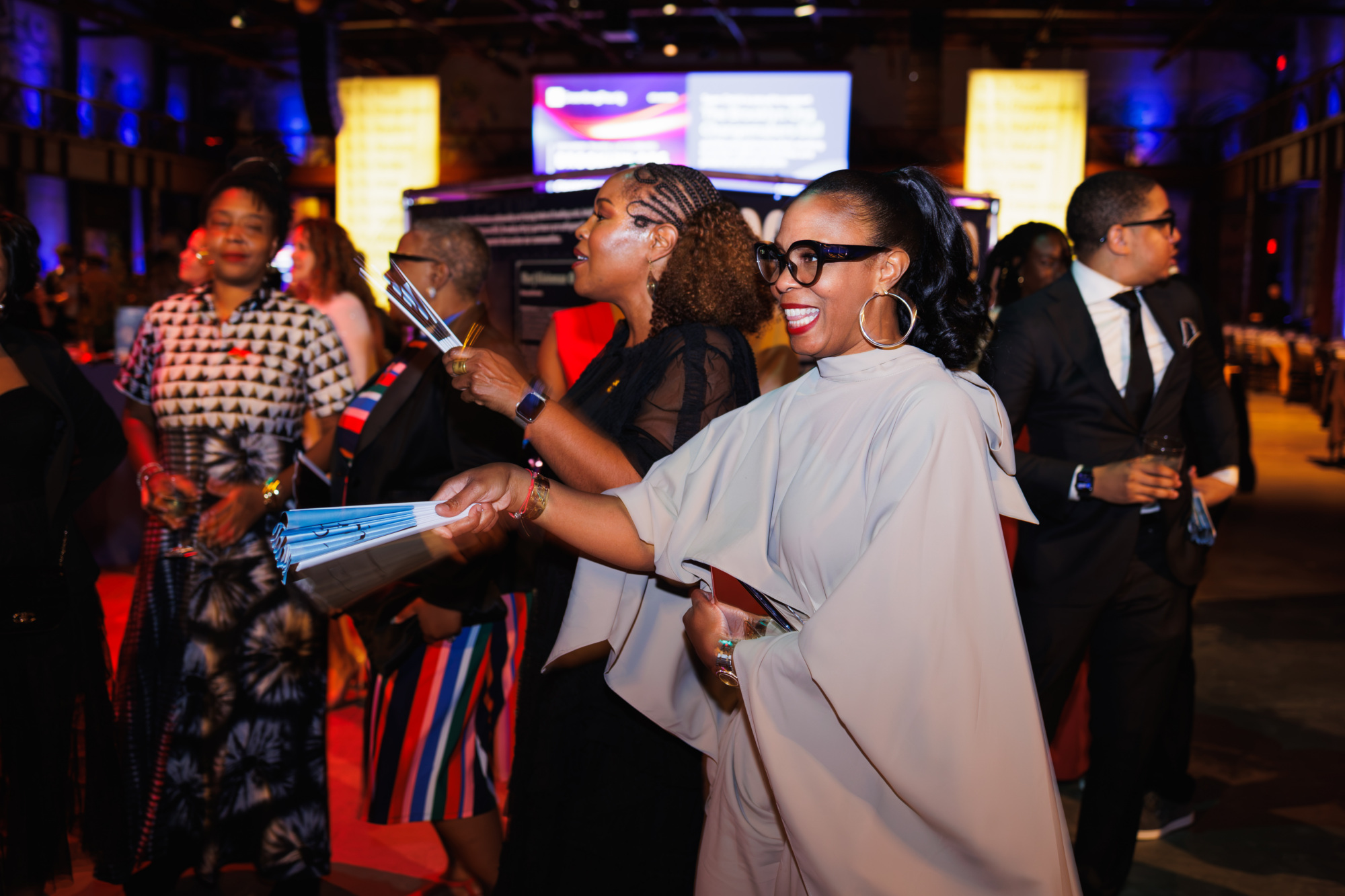 A woman in a cream-colored top and glasses smiles while holding a folded paper fan at a crowded indoor event.