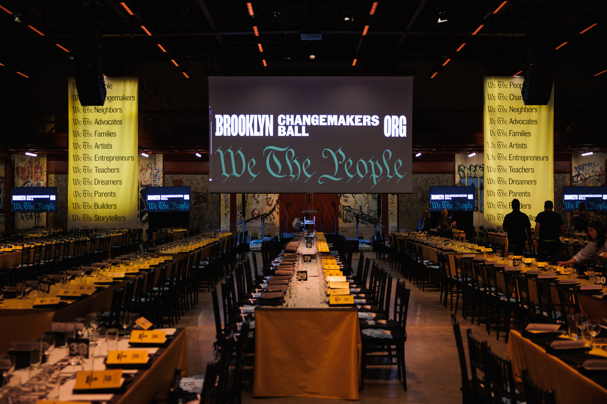 A large hall set for the Brooklyn Changemakers Ball, with a central screen displaying the event name and the phrase “We The People” above long tables with yellow place cards.