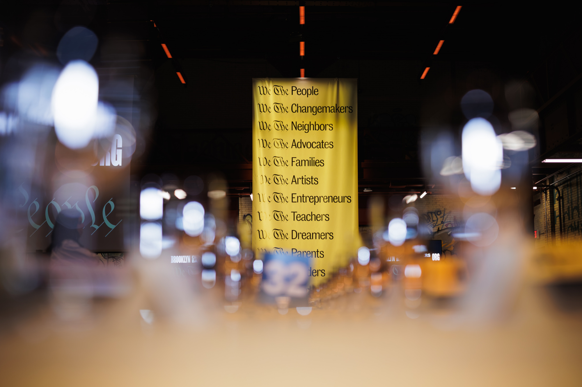 A yellow banner hanging in an indoor space with the heading “We The People” followed by a list of community roles including “Changemakers,” “Neighbors,” “Advocates,” “Families,” and “Artists.”