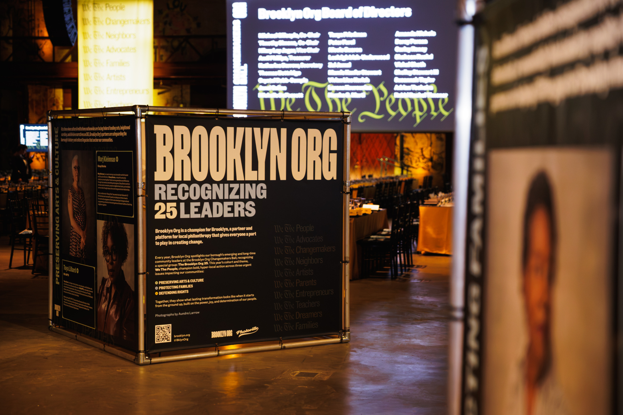 A large square display stand for Brooklyn Org’s “Recognizing 25 Leaders” exhibit, featuring informational text and photos, with a list of the Board of Directors projected on a screen in the background.