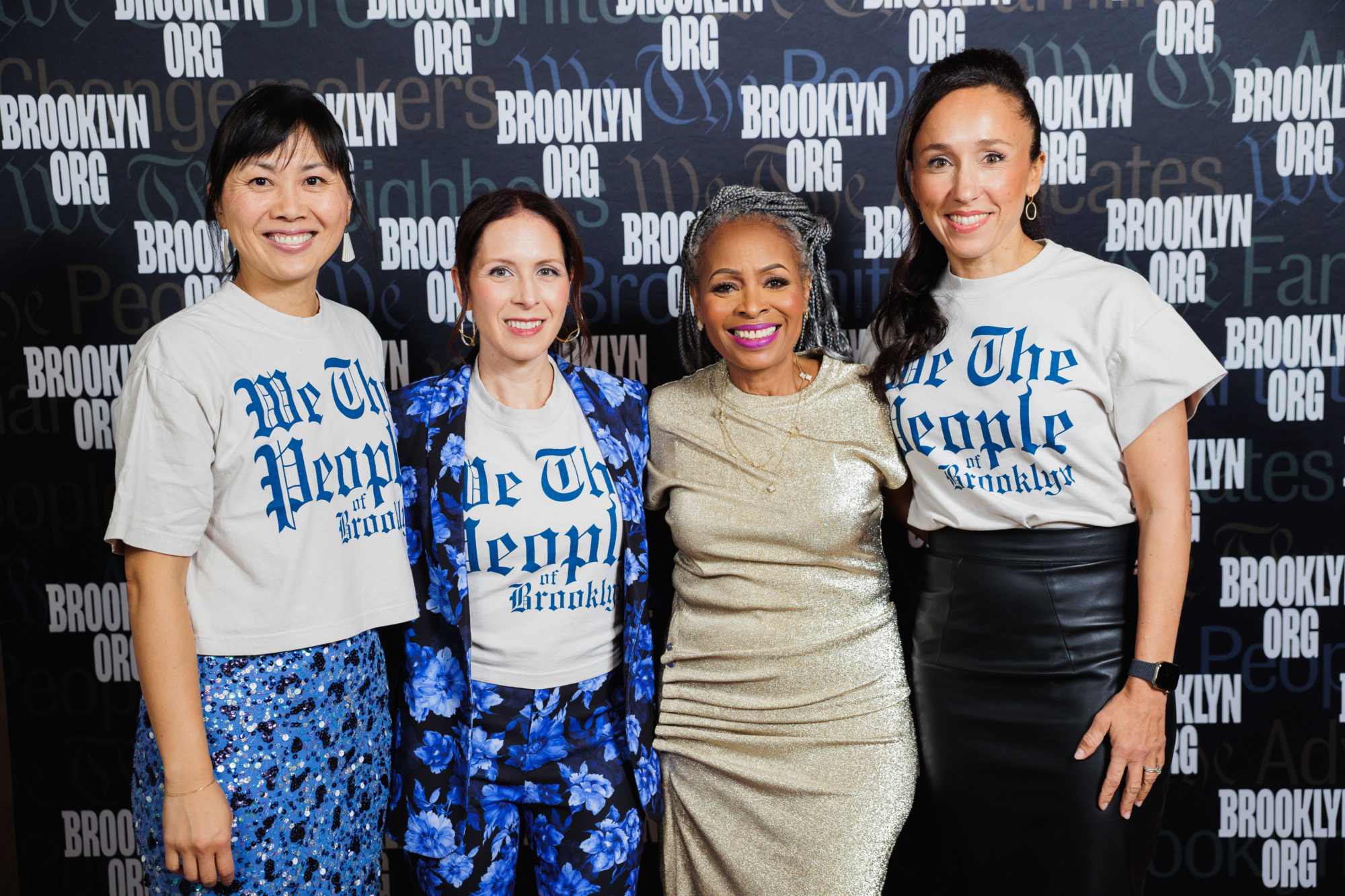 Four women posing in front of a Brooklyn Org backdrop, with three wearing “We The People of Brooklyn” t-shirts.