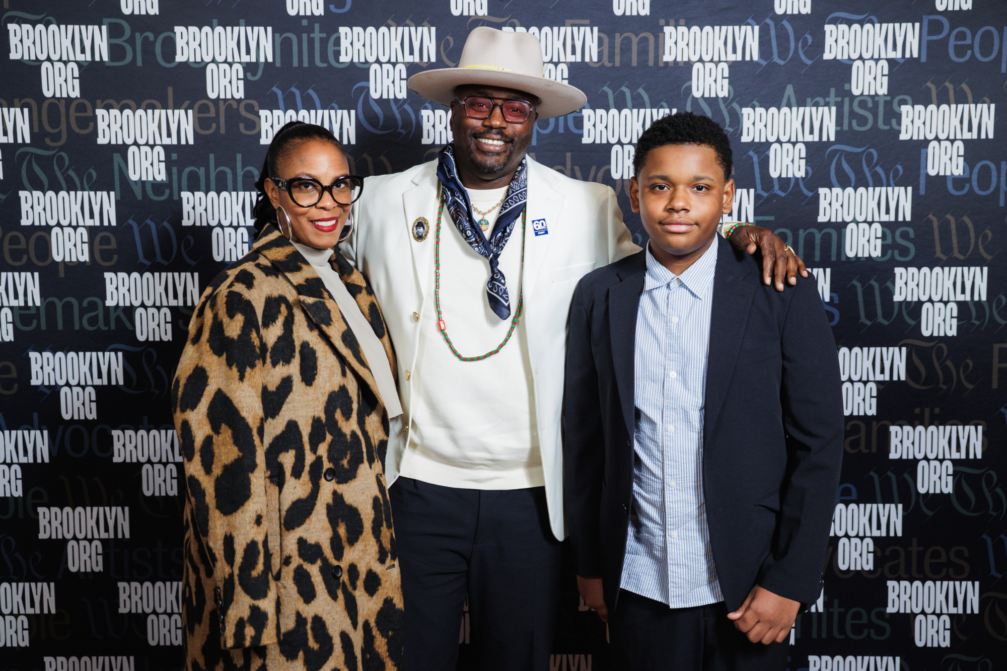 A woman, a man in a hat and blazer, and a boy posing in front of a Brooklyn Org step-and-repeat backdrop.