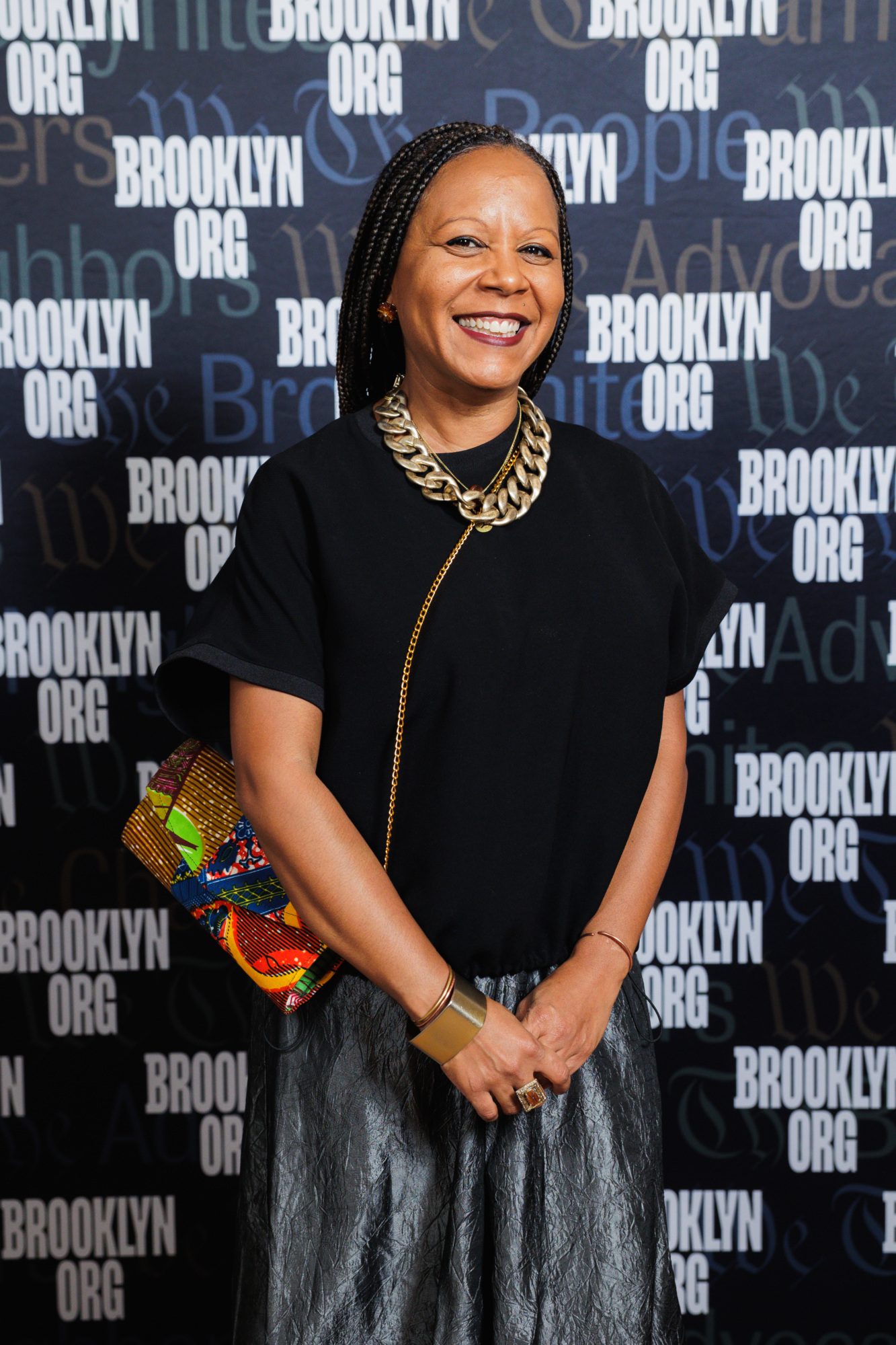 A smiling woman with braided hair wearing a black top, a metallic gold necklace, and a patterned clutch bag, standing in front of a Brooklyn Org step-and-repeat backdrop.