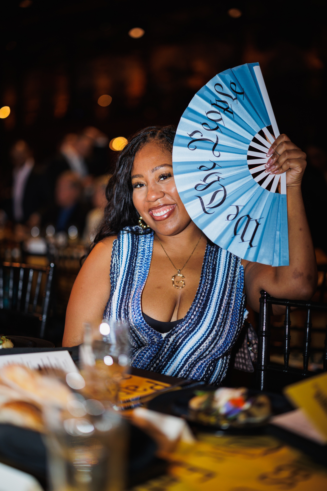 A smiling woman at a table holds up a blue hand fan that says “We the People.”