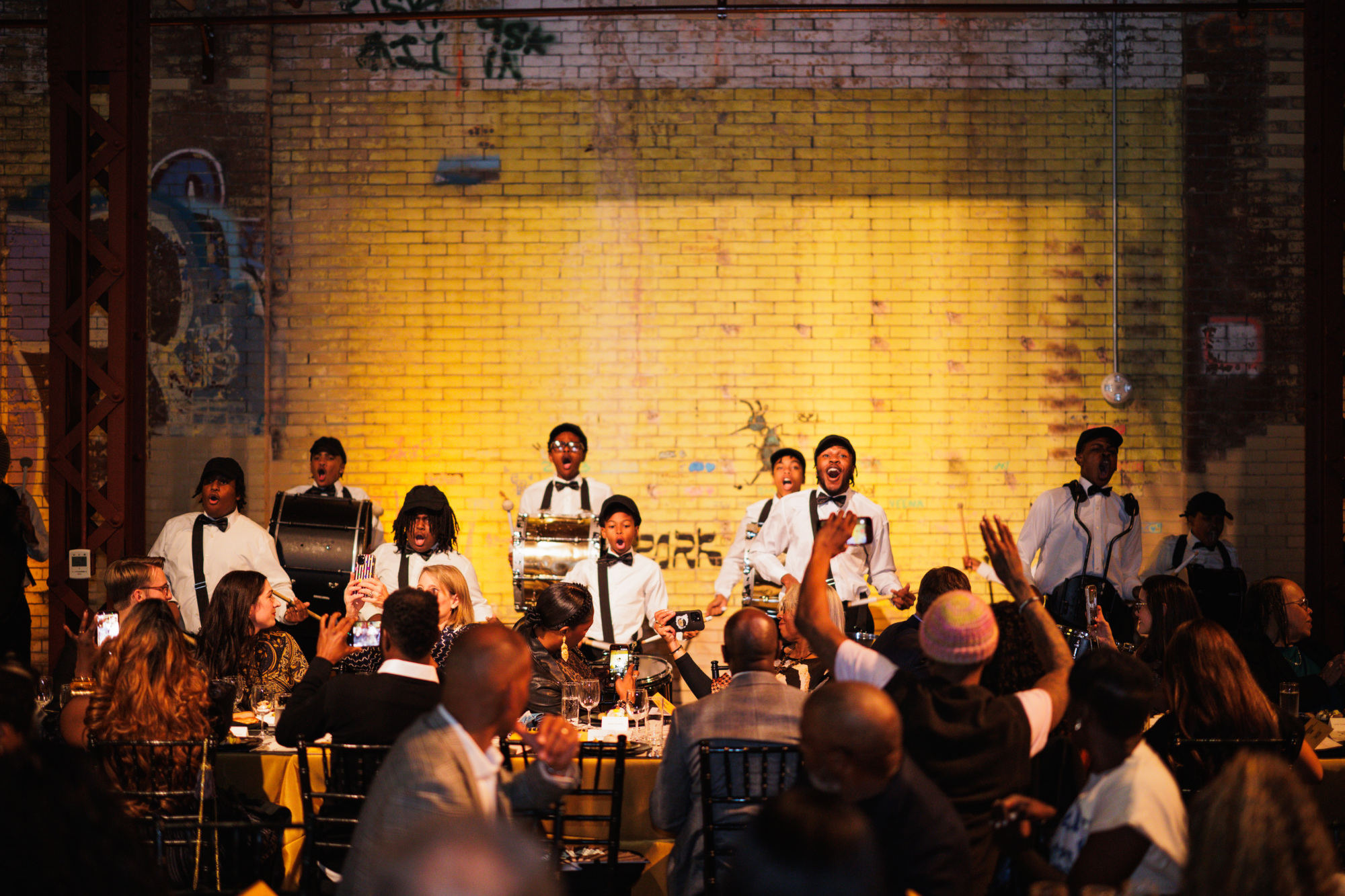 A marching drum line in white shirts and bow ties performs in front of a yellow brick wall for an audience at dinner tables.