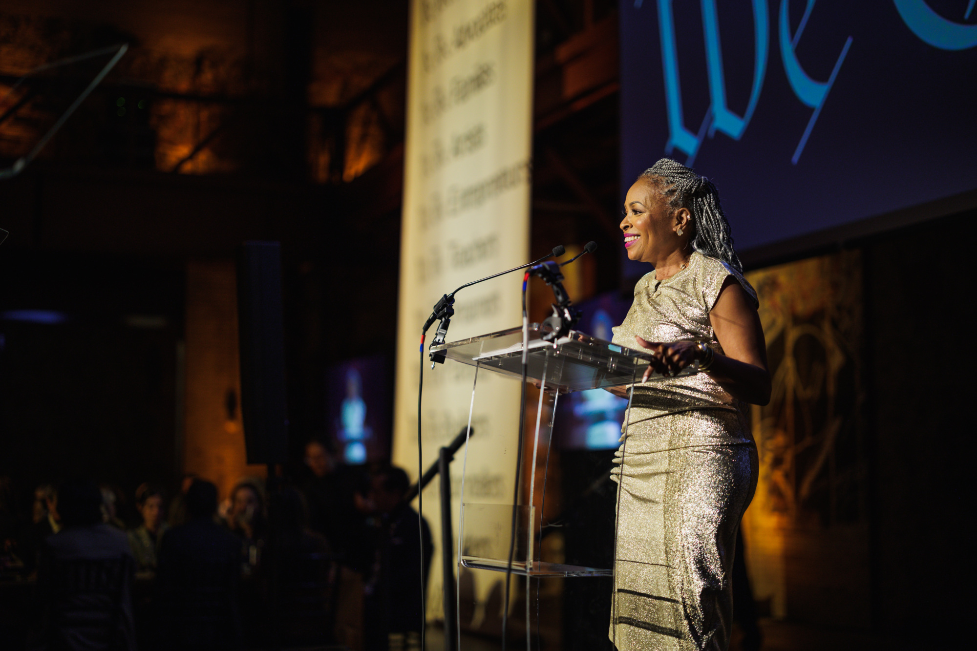 A woman in a gold dress speaking at a podium in a dark room.