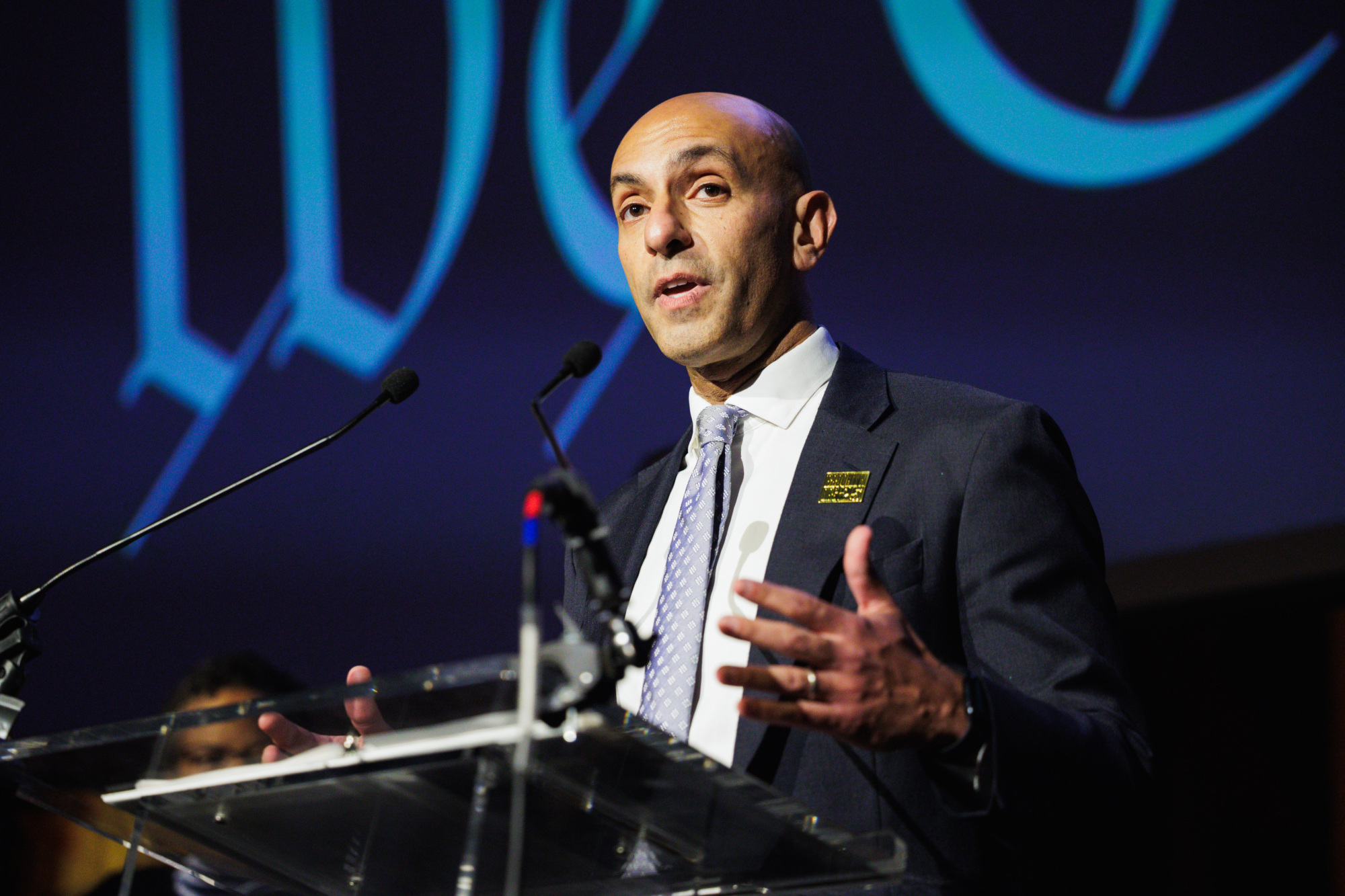 A man in a suit and tie speaks from a podium at an event.