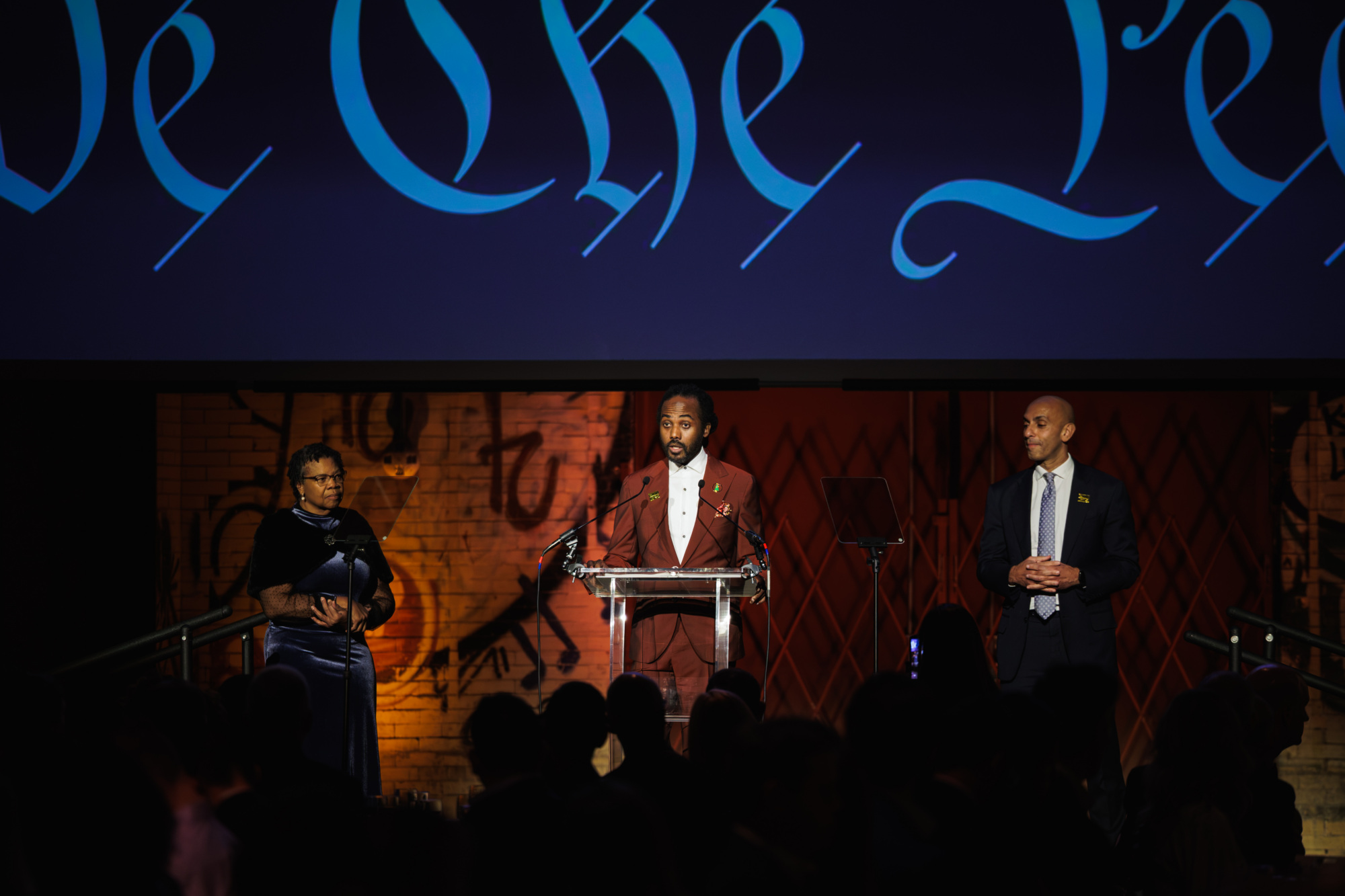 A man in a red suit stands at a podium speaking to an audience, flanked by a woman in a velvet dress and a man in a navy suit, with a large blue banner behind them.