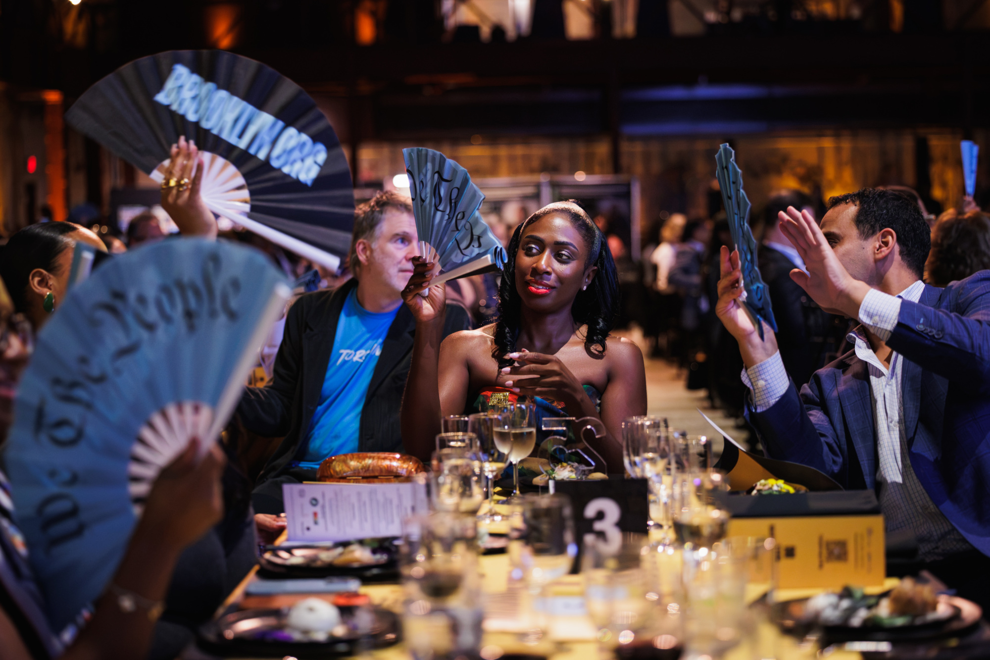 People seated at a dining table holding handheld folding fans with text including “Brooklyn.org” and “We The People.”