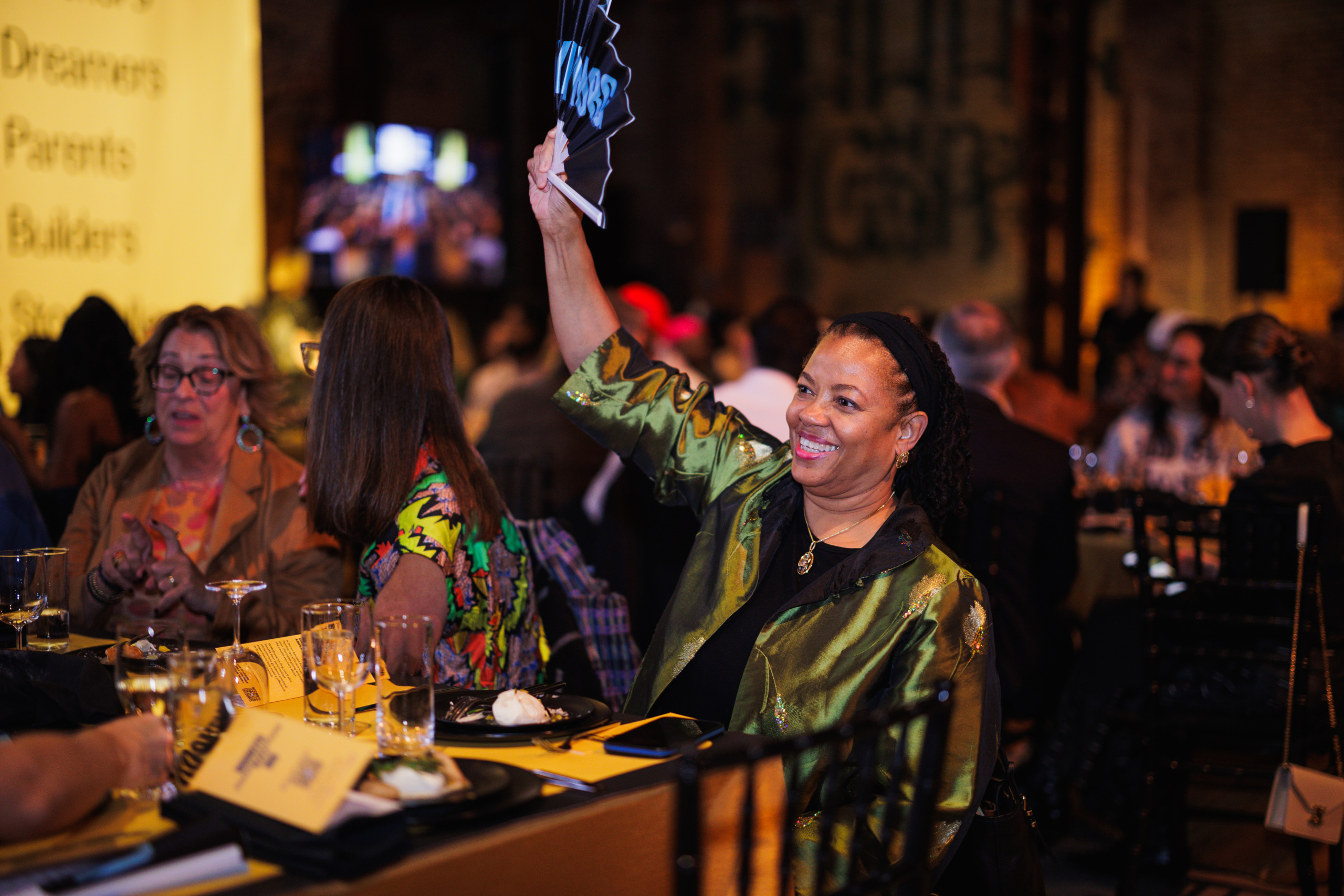 A woman at a banquet table holds up a black hand fan with white lettering.