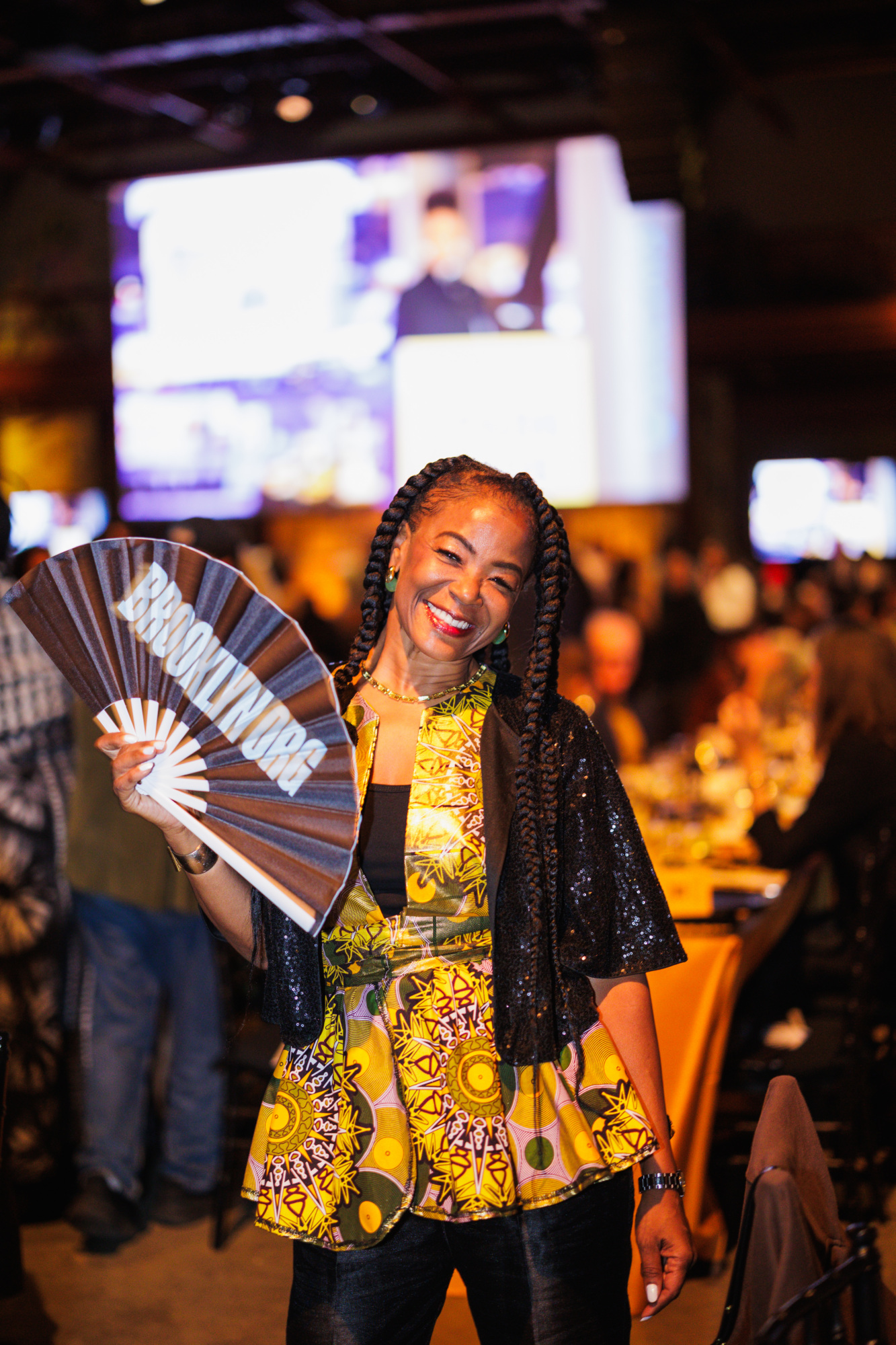 A smiling woman with braided hair holds a folding fan that reads “BROOKLYN.ORG” at an event.