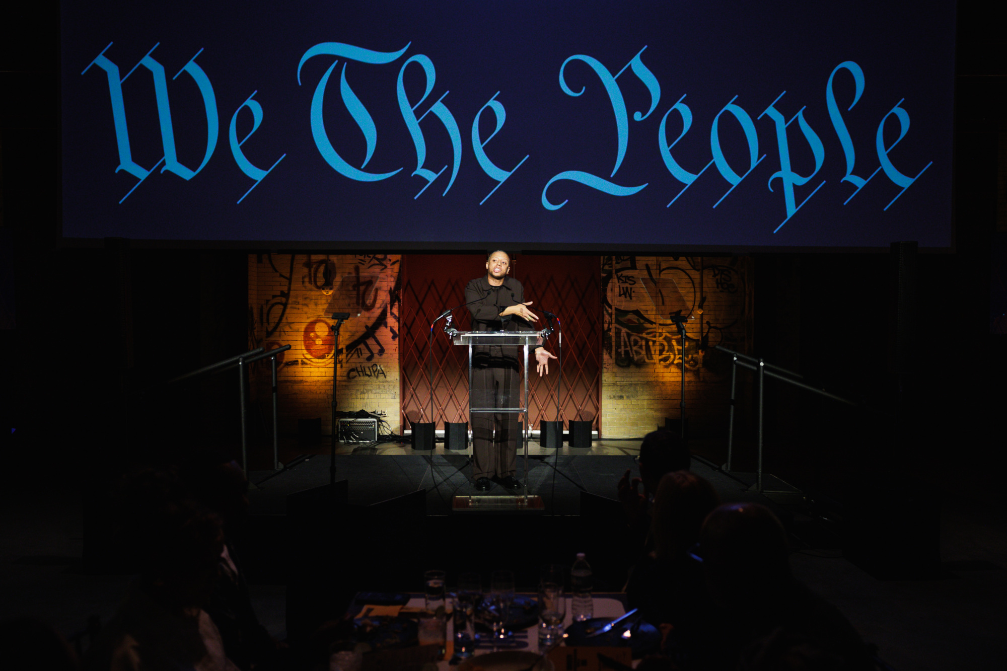 A person speaks at a podium on a stage in front of a large screen displaying the words “We The People.”