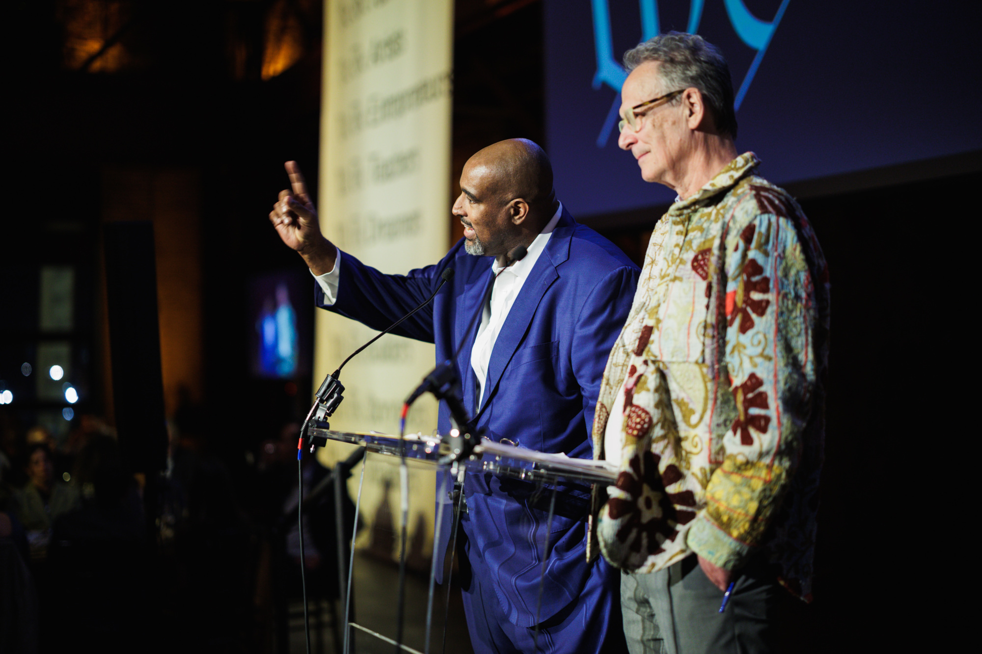 A man in a blue suit speaks at a podium with a raised finger, standing next to a man wearing a patterned shirt.