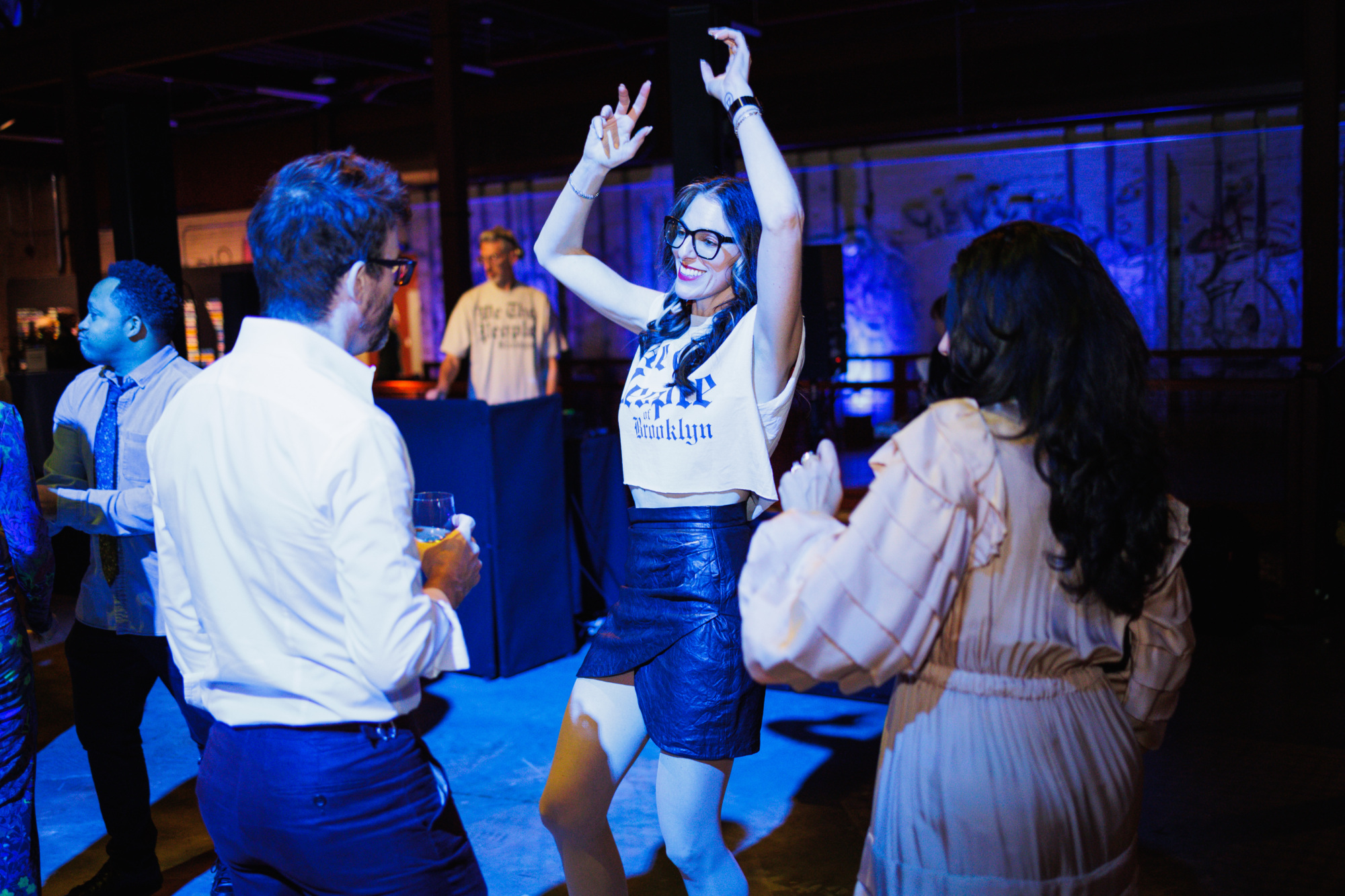 A woman in a white “Brooklyn” crop top and black skirt dances in a dim, blue-lit room while others look on.