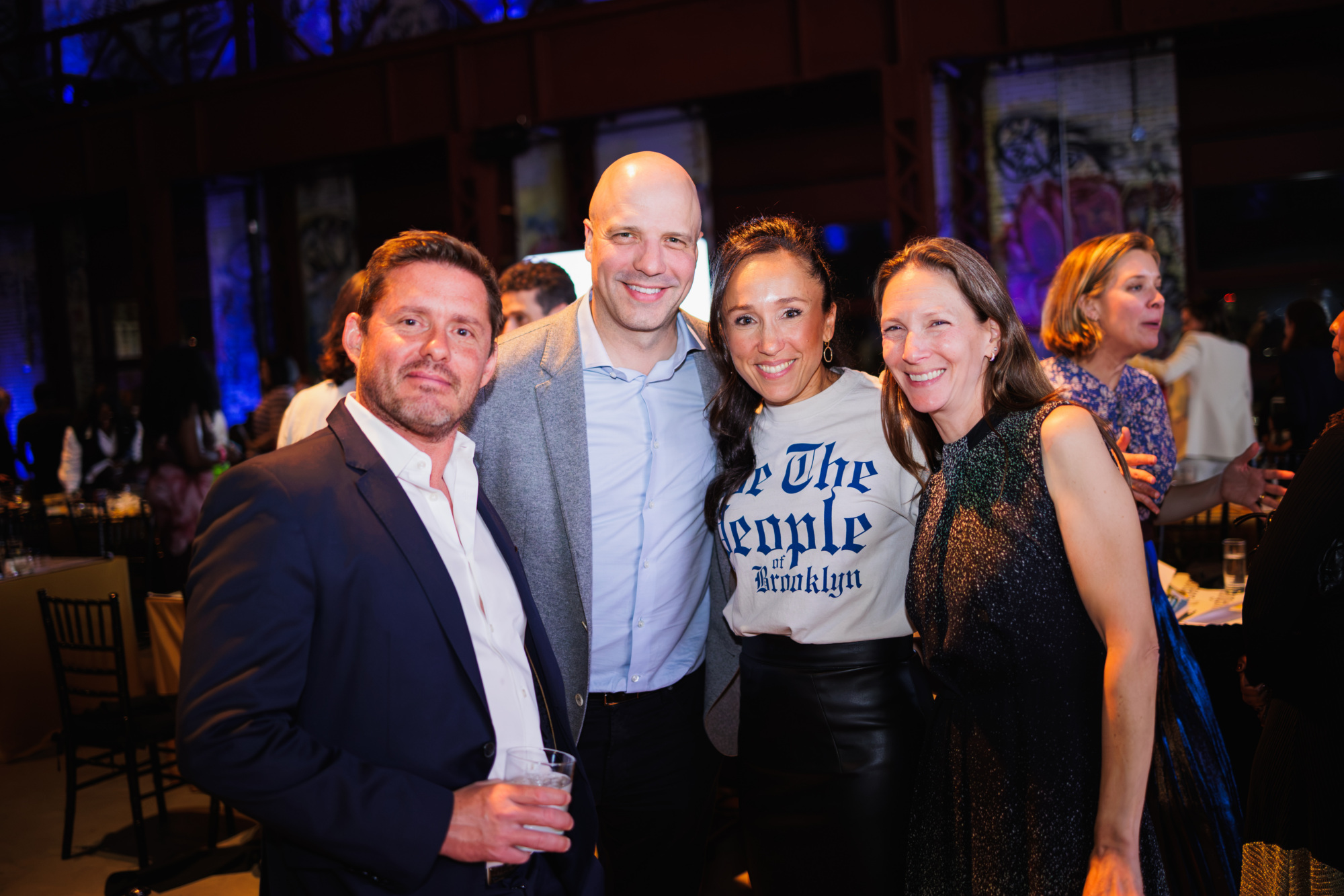 Four people posing together at an indoor event, including a woman wearing a shirt that says “We The People of Brooklyn.”