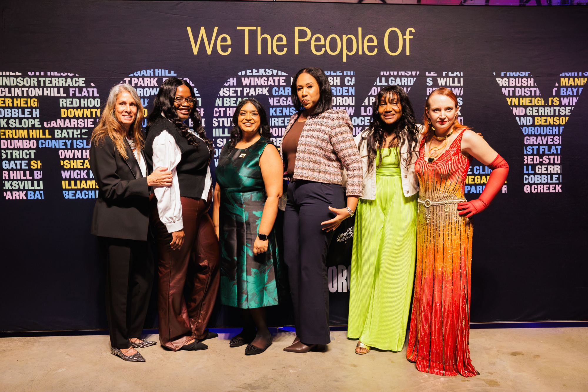 Six women posing together in front of a black backdrop featuring the words “We The People Of” and a list of New York City neighborhoods.