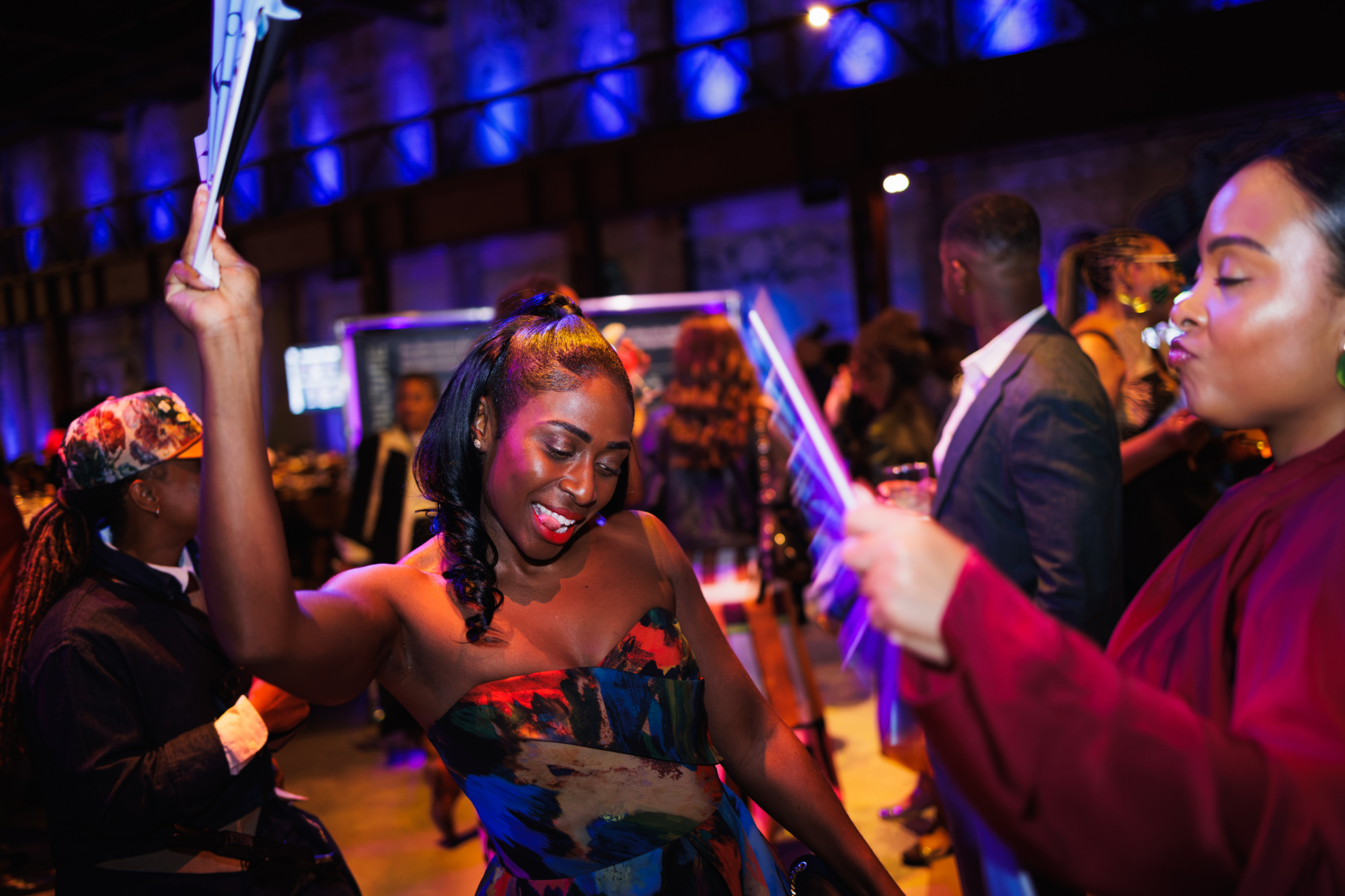 A woman in a colorful strapless dress dances at an indoor event, holding a fan overhead.