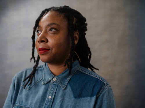 A person with locs and red lipstick looks off to the side, wearing a denim shirt and triangle earrings.