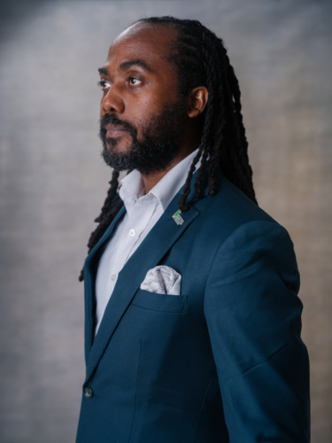 A man with locs and a beard wearing a white dress shirt and a teal blazer with a patterned pocket square.