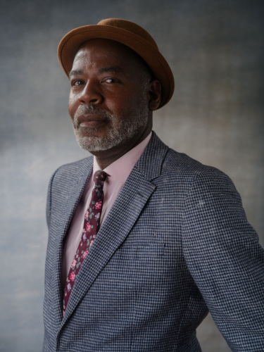 A middle-aged man with a graying beard, wearing a brown fedora, a plaid blazer, a pink dress shirt, and a floral tie.