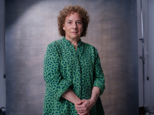 A woman with curly hair wearing a green shirt with a leaf pattern, standing against a grey backdrop.