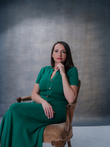 A woman in a green dress sitting in a chair with her hand to her chin.