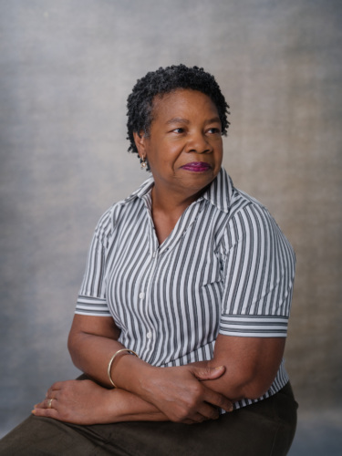 A woman with short dark hair wearing a short-sleeved, grey and white striped collared shirt sits with her arms crossed.