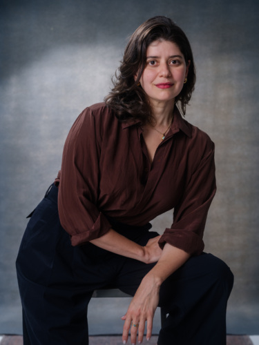 A woman with shoulder-length brown hair wears a brown button-down shirt and black trousers, looking toward the camera.