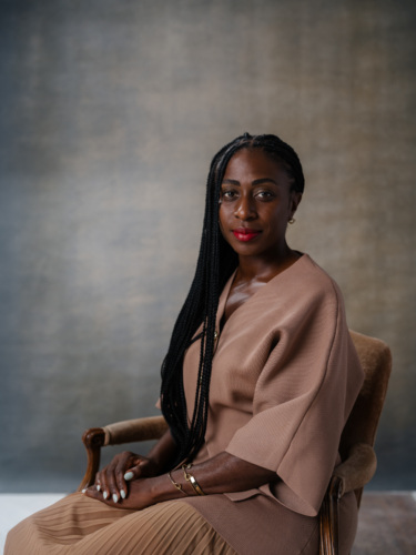 A woman with braided hair and red lipstick sitting in a chair, wearing a beige top and pleated skirt.