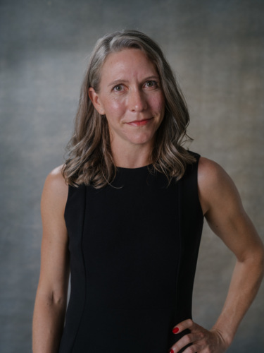A woman with shoulder-length grey-brown hair wearing a black sleeveless dress, posing against a grey backdrop.