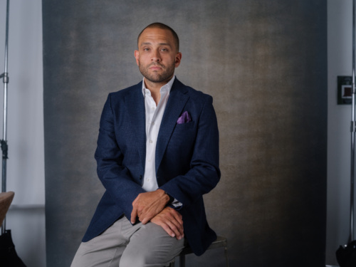 A man in a navy blazer, white shirt, and grey trousers sits on a stool against a grey backdrop.